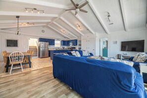 A spacious living area with a large blue sofa, wooden dining table, and chairs. A kitchen with blue cabinets and a refrigerator is visible in the background. - The Sailfish - Duck, Carolina Dunes, Beach 300 Yds, Sound View, Pool, Hot Tub, Pets OK, Flex Stays (Duck)