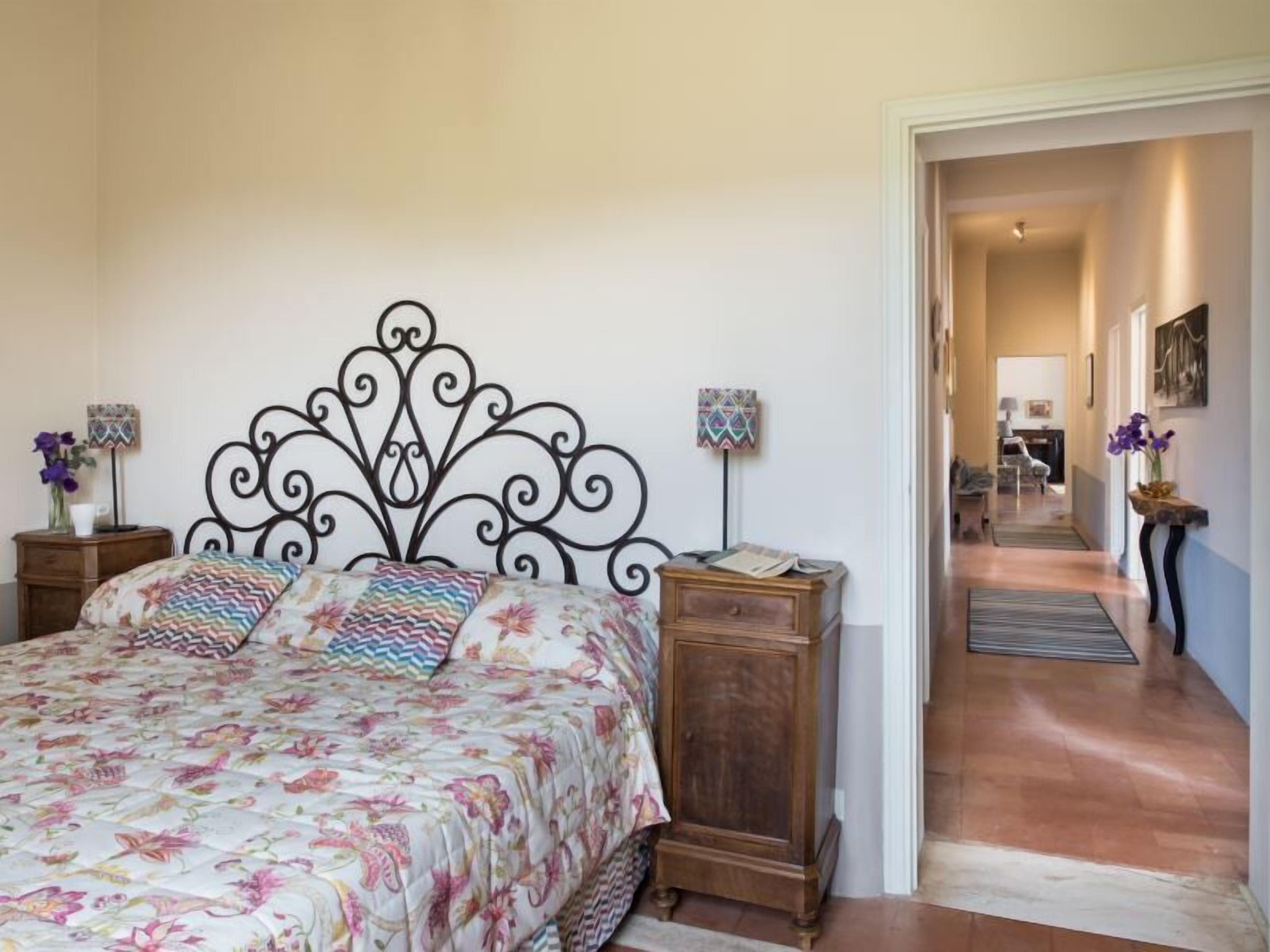 A bedroom with a floral bedspread, a decorative headboard, bedside tables with lamps, and a view into the living area.