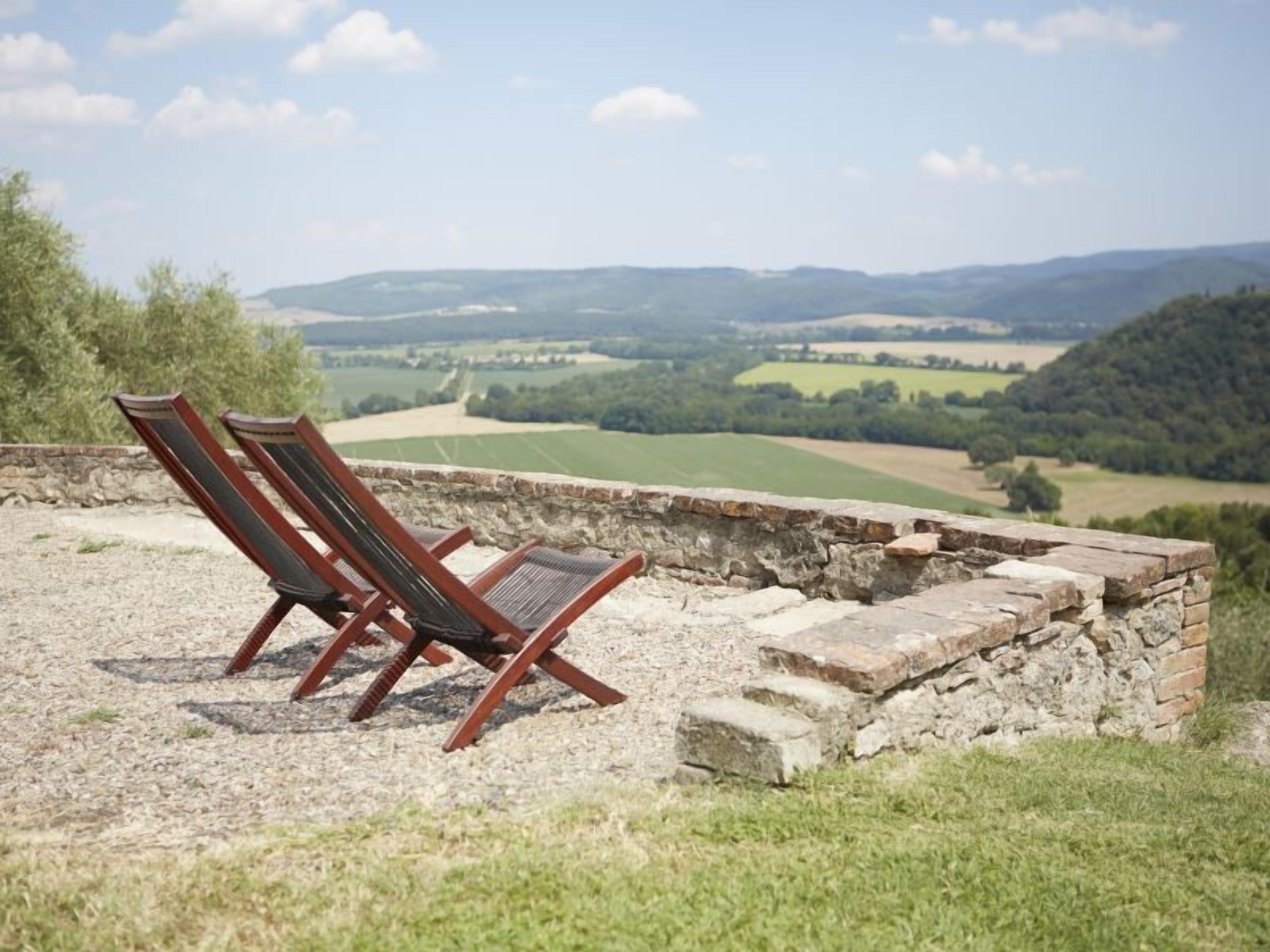Un mur de pierre bordé de deux fauteuils en bois, placés de manière à offrir une vue panoramique sur des collines et des champs vallonnés.