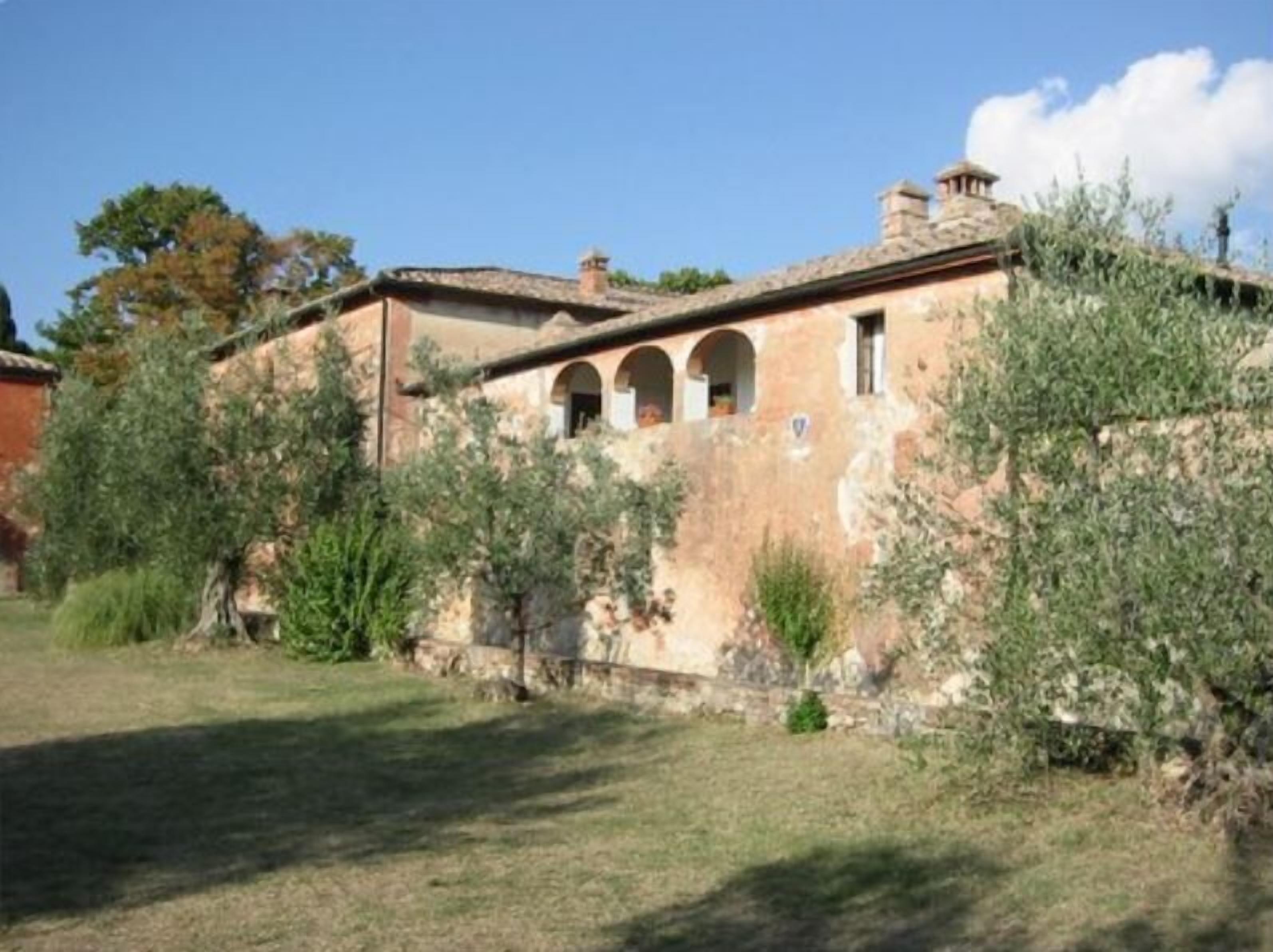 A rustic stone building with a tiled roof, arched windows, and a small garden area with olive trees.