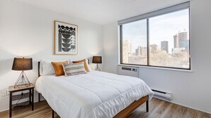 Un dormitorio moderno con un ventanal grande, una cama con ropa blanca, dos lámparas de noche y vistas a la ciudad.