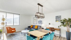 Una sala de estar moderna con una mesa de comedor, sillas color turquesa, un sofá con cojines amarillos y vistas a la ciudad a través de amplios ventanales.