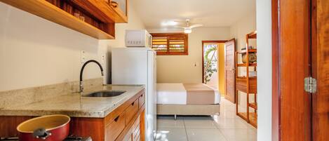 A compact kitchen with a stove, sink, and microwave. A bed is visible in the background, and a wooden door is on the right.