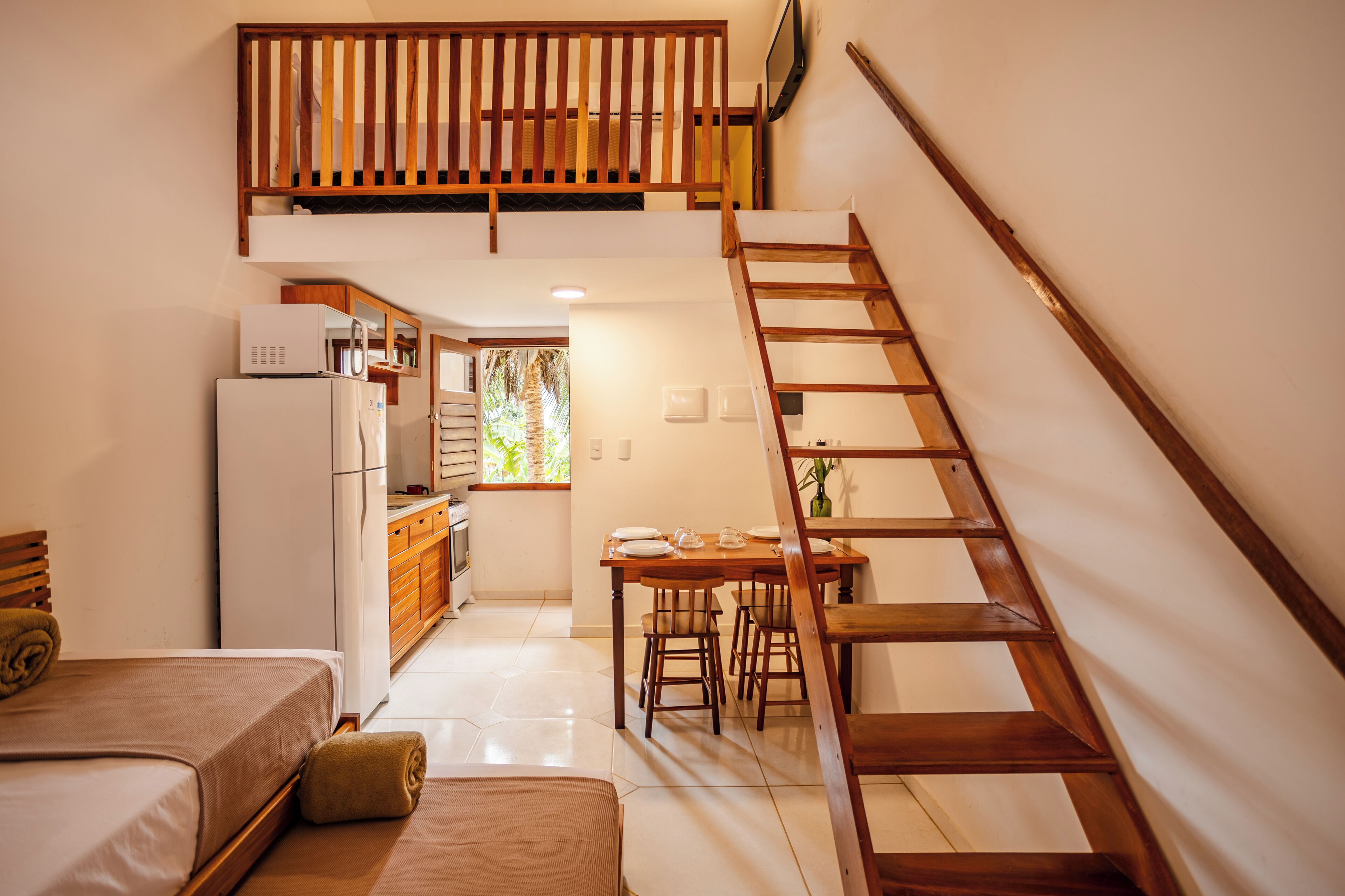 A compact hotel room with a bed, a small kitchenette, a dining area with a table and chairs, and a wooden staircase.
