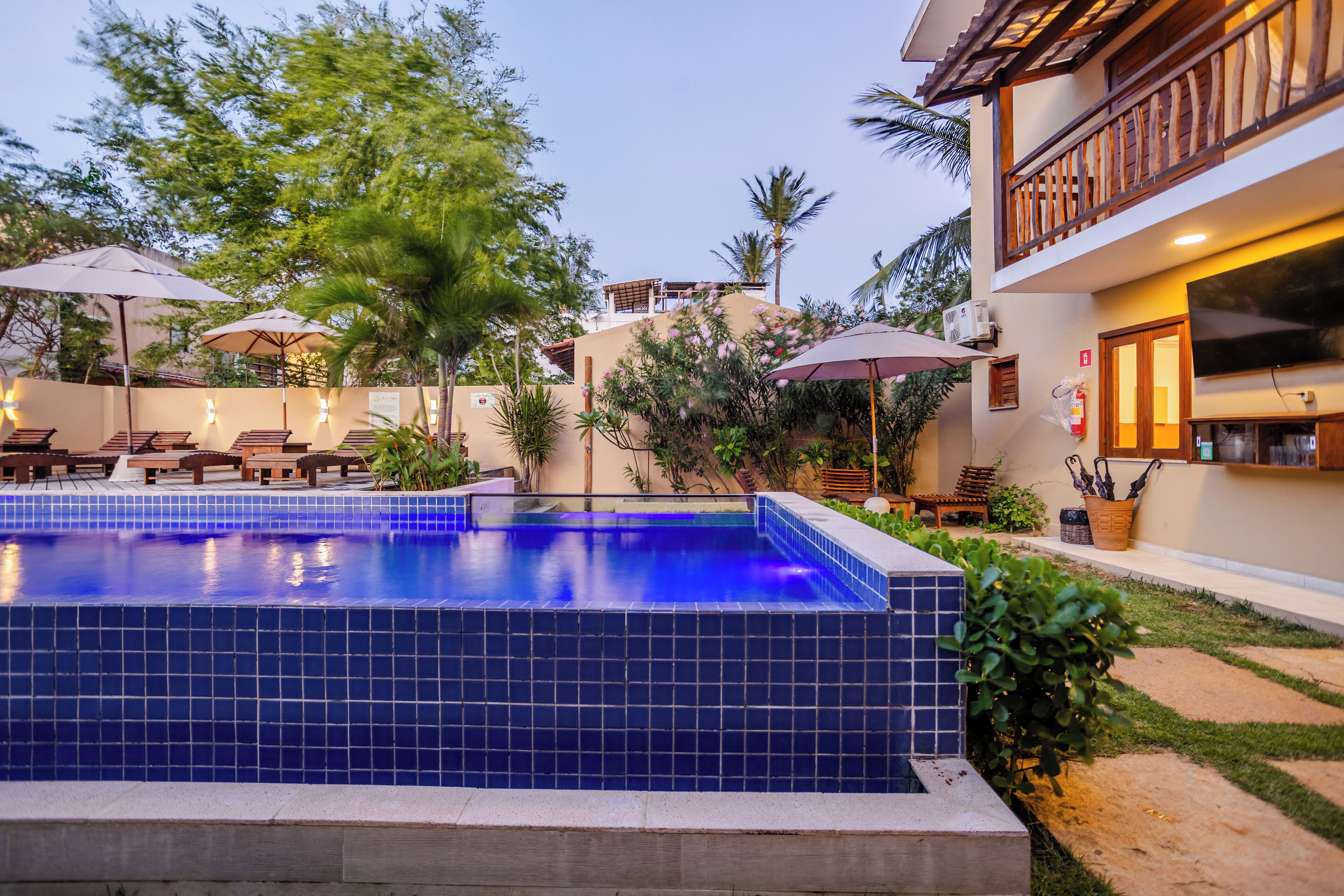 A swimming pool with lounge chairs and umbrellas, surrounded by greenery and a building with a balcony.