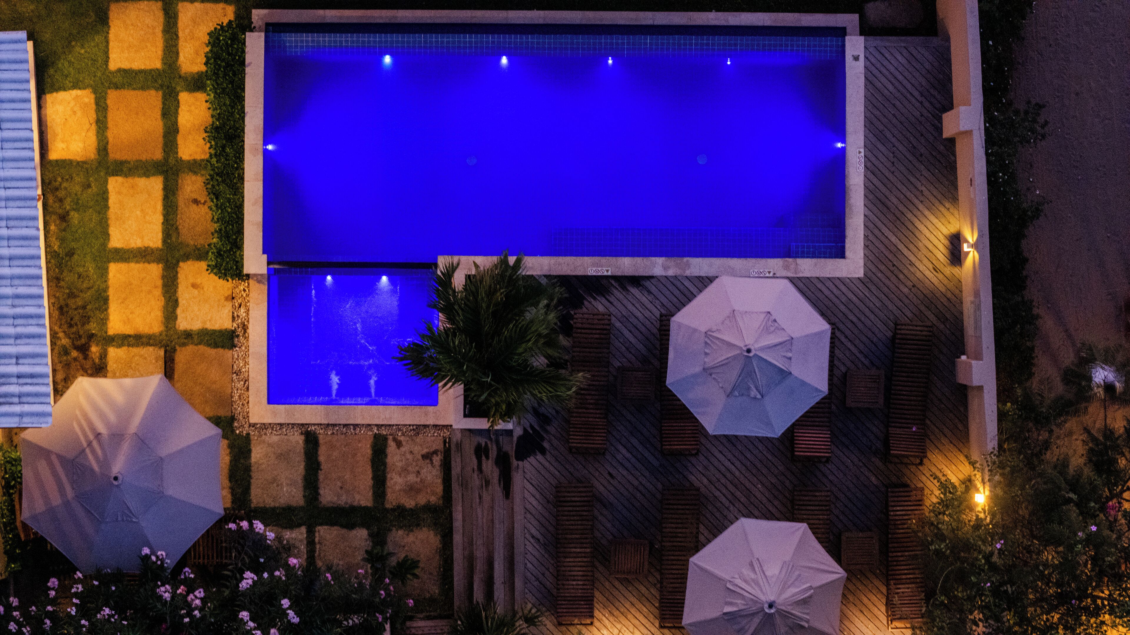 Vista aérea de una piscina rectangular con dos piscinas más pequeñas, rodeada por un jardín con sombrillas y una valla de madera.