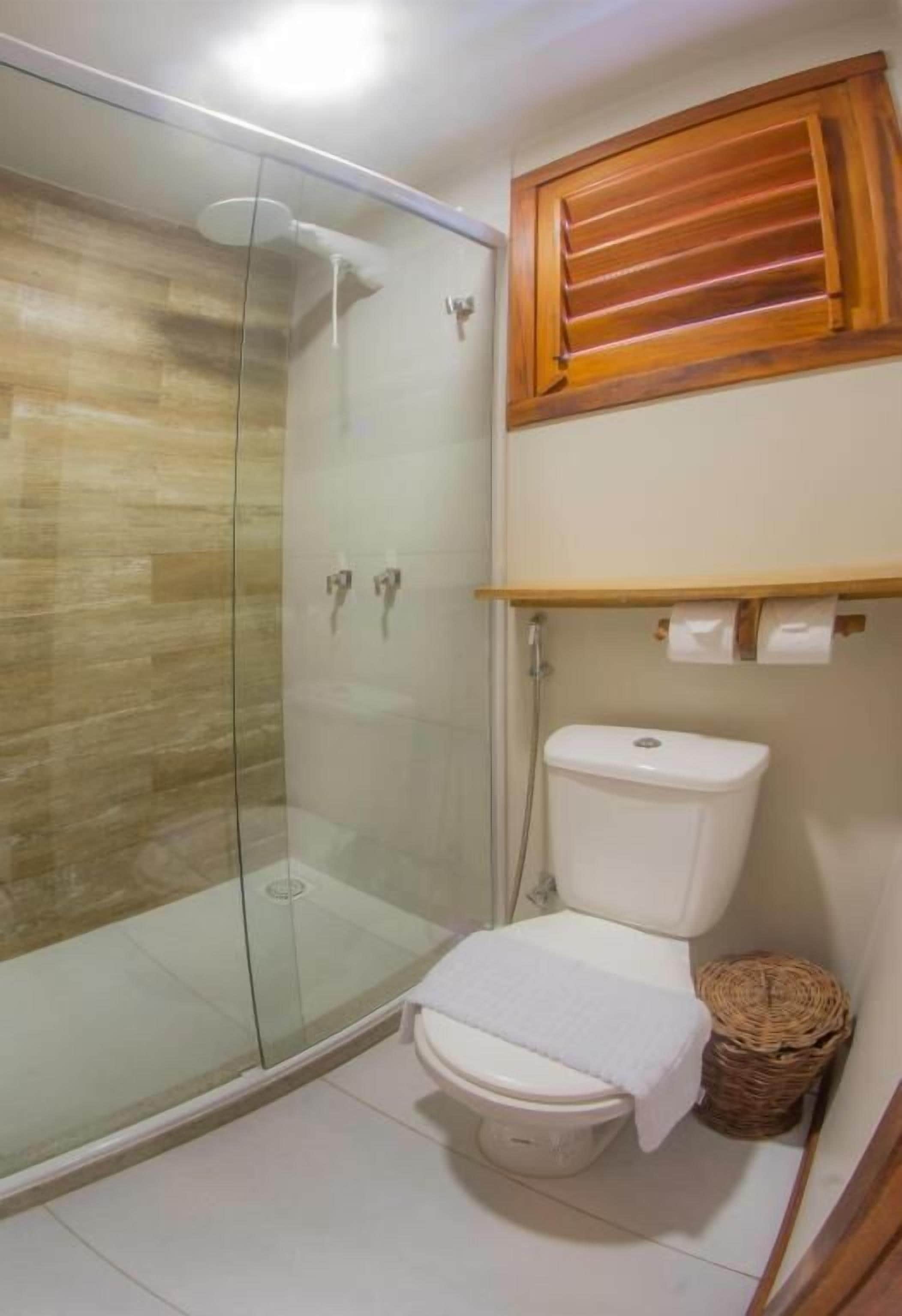 Un baño con ducha envuelta en vidrio, un inodoro blanco y una ventana de madera sobre el inodoro.