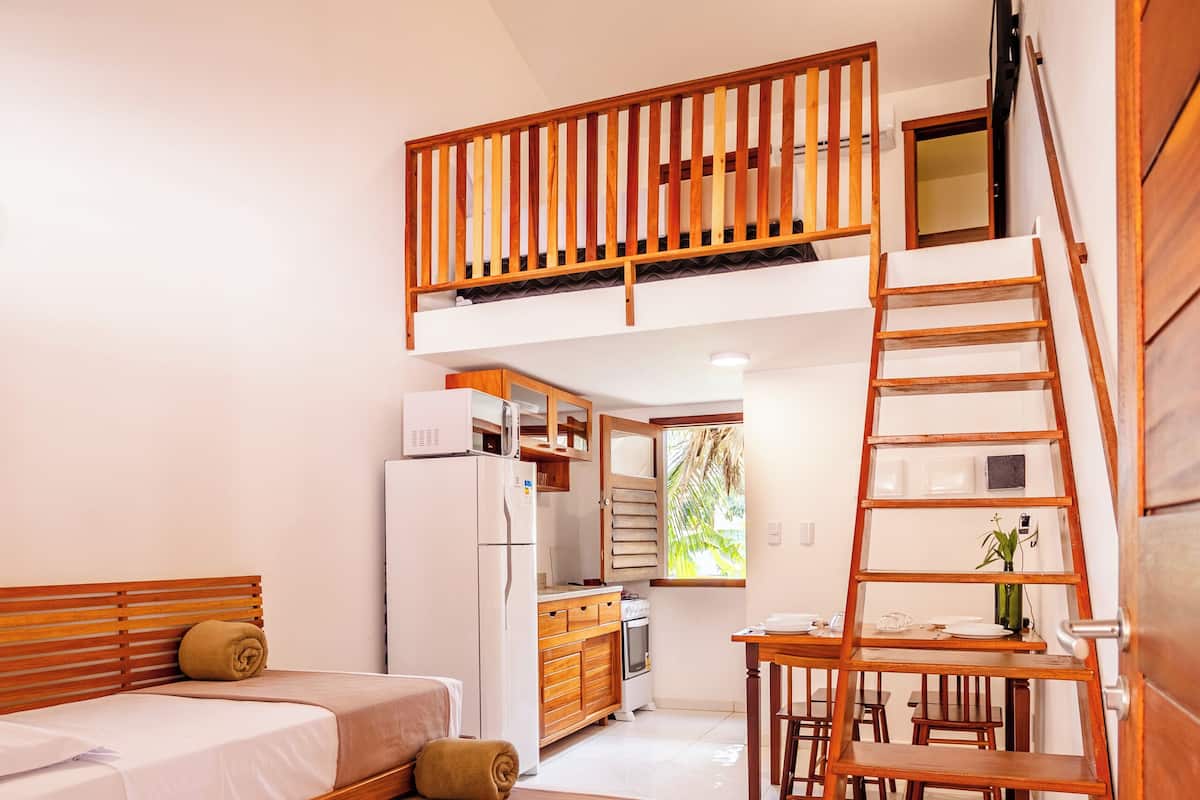A compact, modern room with a loft bed, a small kitchenette, and a wooden staircase.