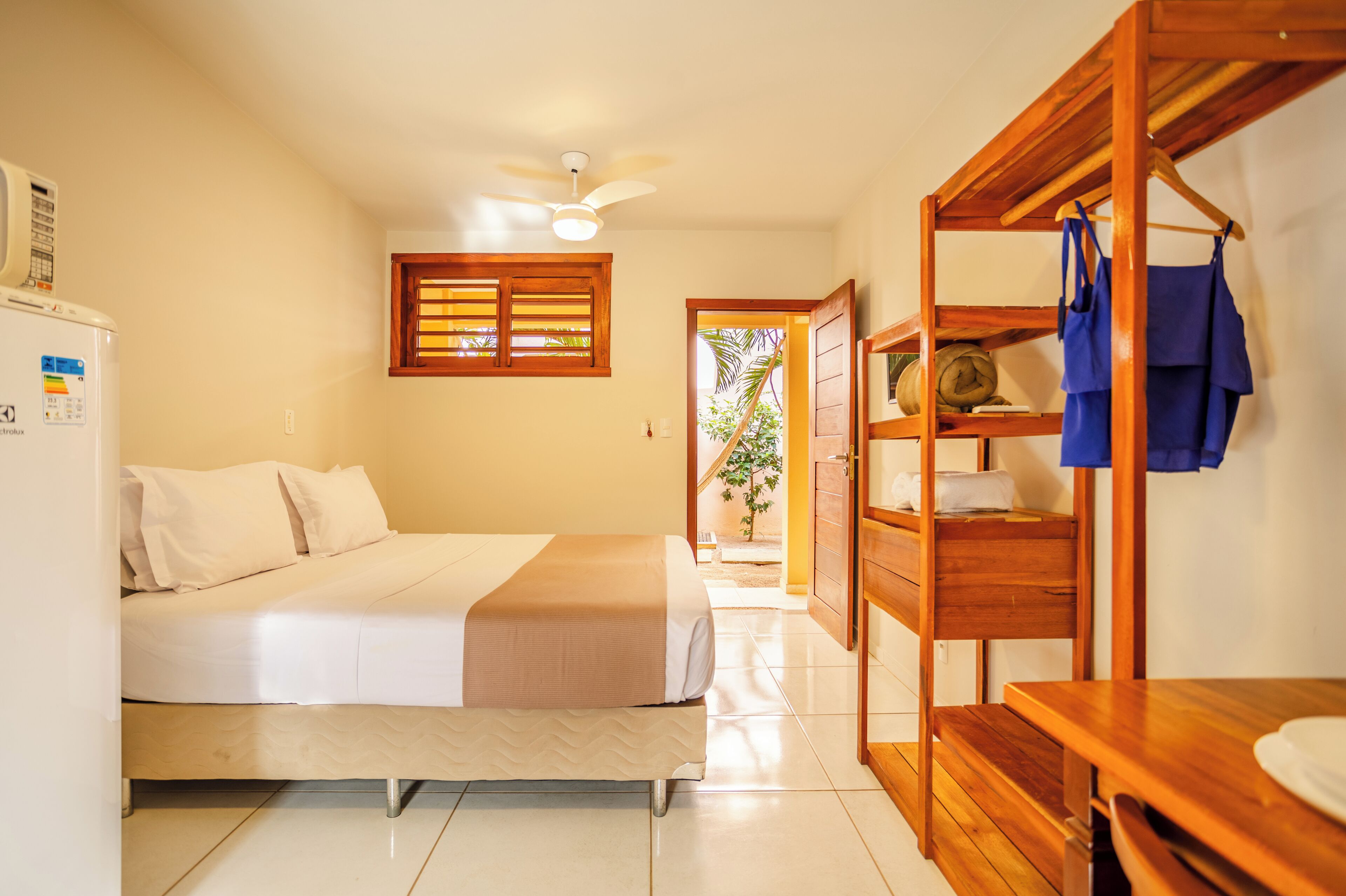 Een hotelkamer met een bed, een houten tafel, een bankje, een kledingkast, een plafondventilator en een raam met jaloezieën.