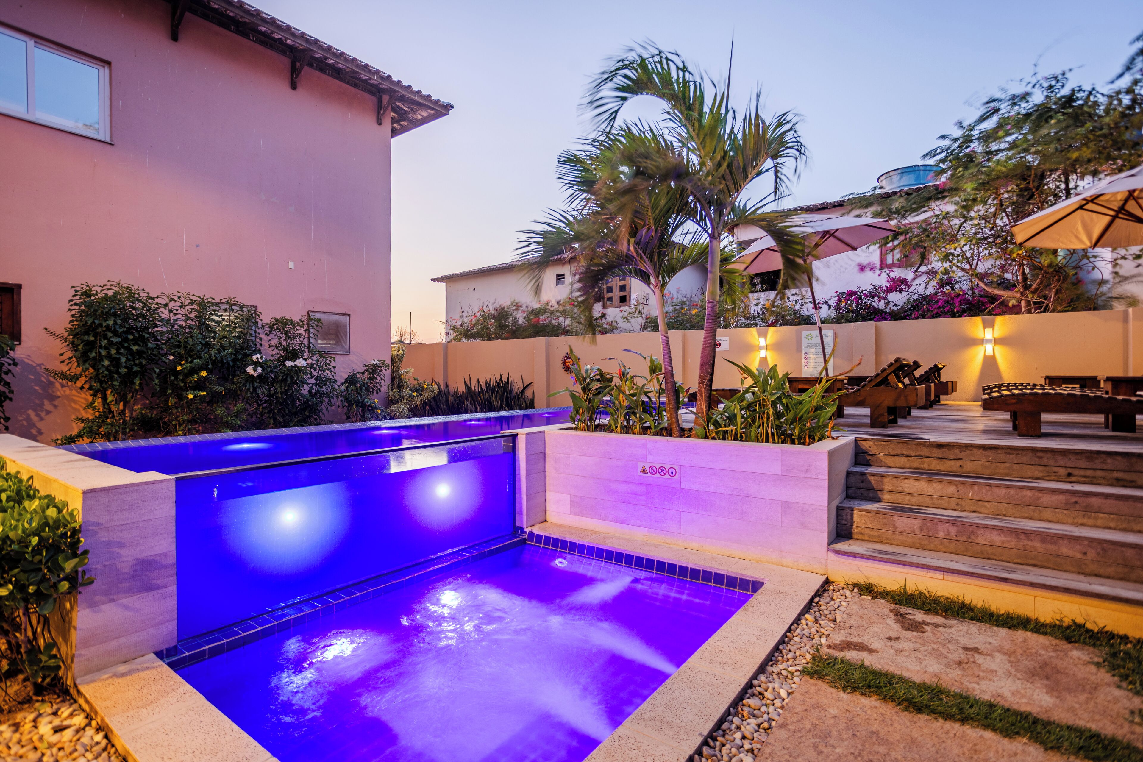 A swimming pool with a blue-lit interior, surrounded by a landscaped garden with palm trees and a patio area with outdoor seating.