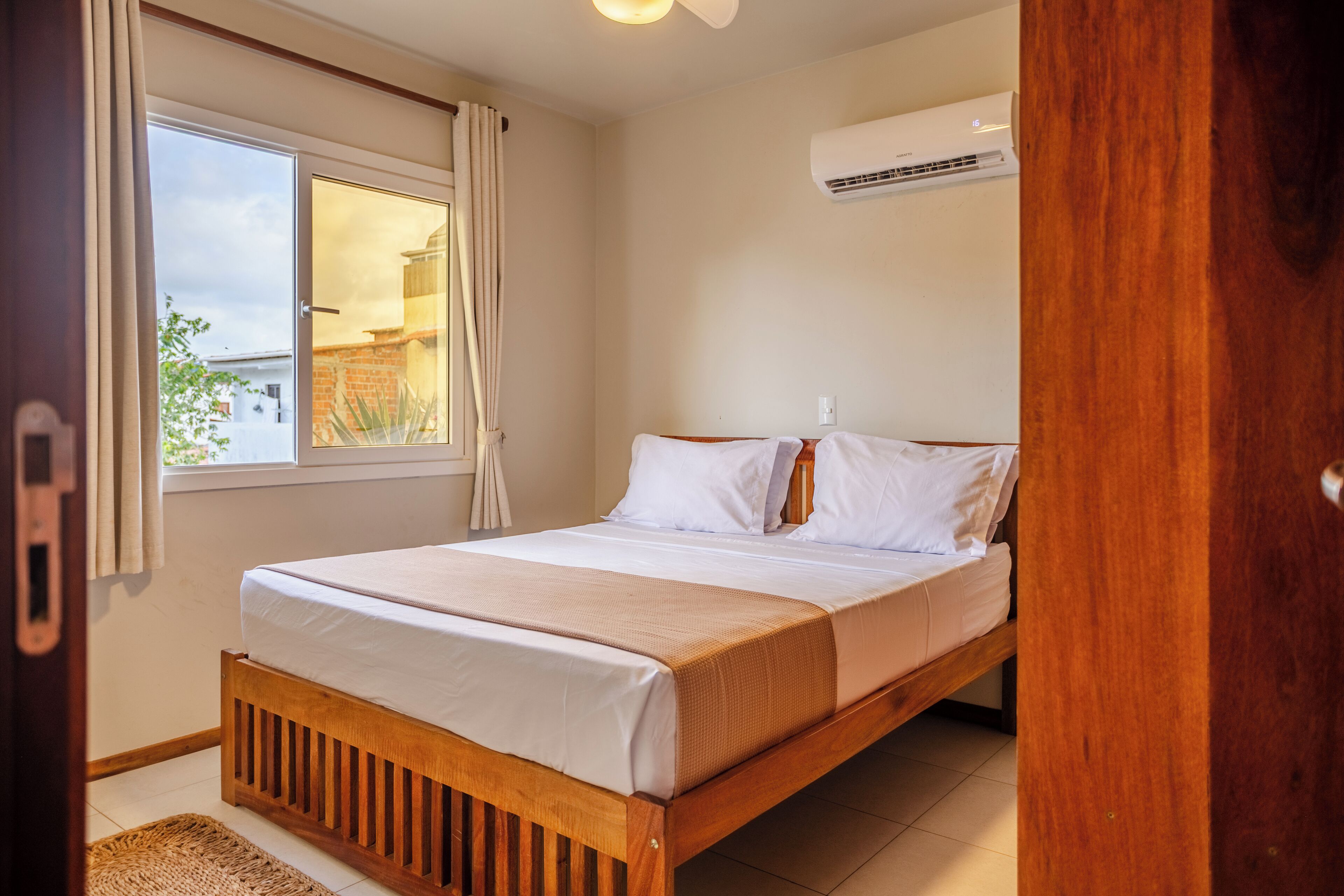 Un dormitorio con cama de madera, ropa de cama blanca, una ventana con cortinas y un equipo de aire acondicionado en la pared.