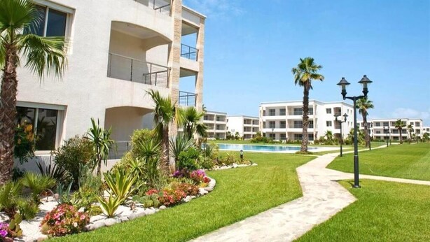Un hotel de varios pisos con piscina, rodeado de césped cuidado y palmeras.