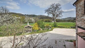 Paesaggio rurale con un edificio in pietra, un albero grande e un sentiero sterrato che conduce a un campo verde.