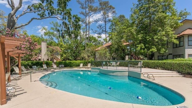 A swimming pool with lounge chairs and a wooden pergola, surrounded by trees and a building. - Desirable Ground Floor Condo in a Gated Community with Pool & Fitness Center! (Saint Simons Island)