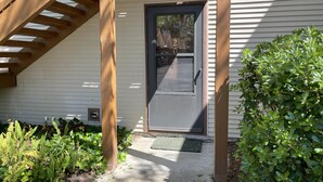 A wooden porch with a door and a small window, surrounded by greenery and a concrete path. - Desirable Ground Floor Condo in a Gated Community with Pool & Fitness Center! (Saint Simons Island)