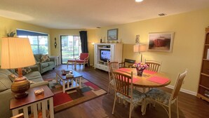 A living room with a sofa, a coffee table, a dining table with chairs, a TV, and a bookshelf. - Desirable Ground Floor Condo in a Gated Community with Pool & Fitness Center! (Saint Simons Island)