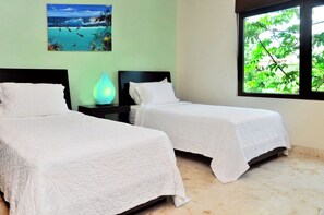 A hotel room with two beds, a nightstand with a lamp, a painting of a beach scene, and a window with a view of greenery.