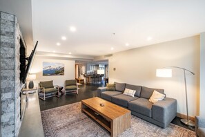 A modern living room with a grey sofa, a wooden coffee table, and a TV mounted on a stone wall. - Blackcomb Ski-in/out | Private hot tub | BBQ (Whistler)
