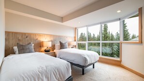 Una habitación de hotel moderna con dos camas, ventanas amplias que dan a una vista de bosque y un cabecero de madera.