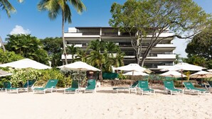Ein Strand mit Liegestühlen und Sonnenschirmen, Palmen und ein mehrstöckiges Gebäude im Hintergrund.