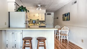 Een keuken met een centraal eiland, twee houten krukken, een eettafel met stoelen en een plafondventilator.