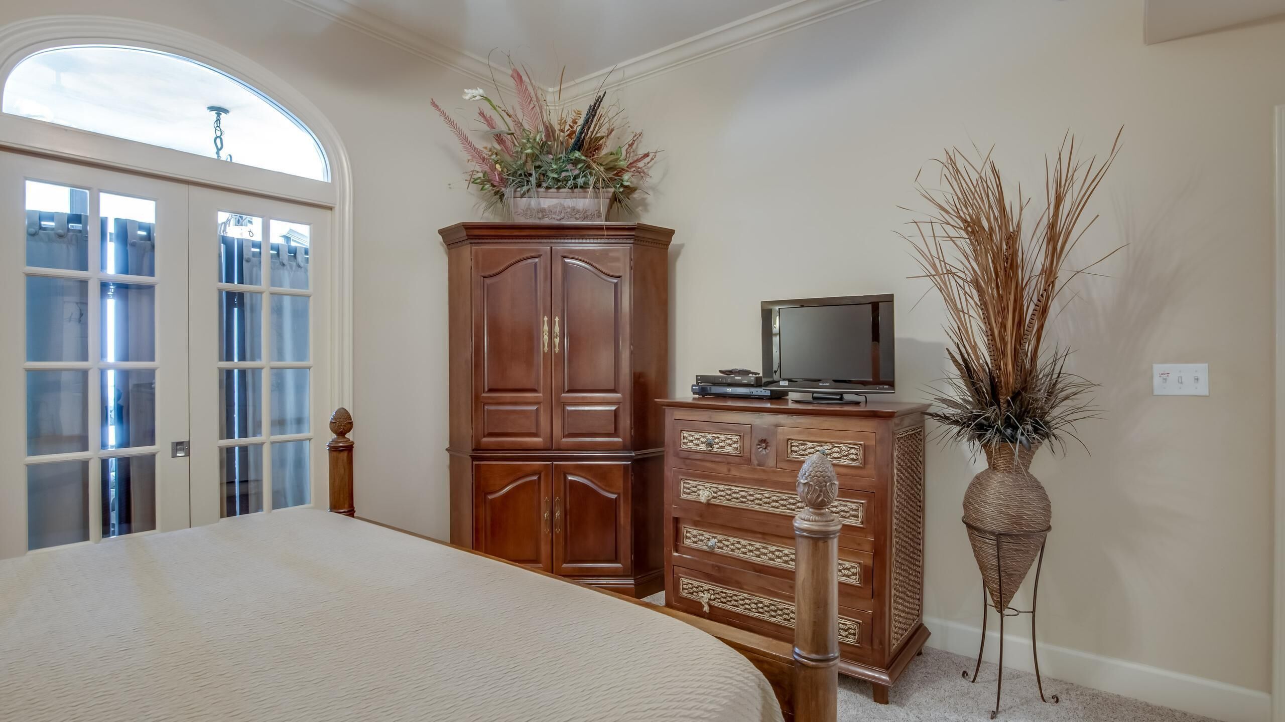 A bedroom with a large bed, a wooden wardrobe, a television on a stand, and a decorative vase with dried plants.