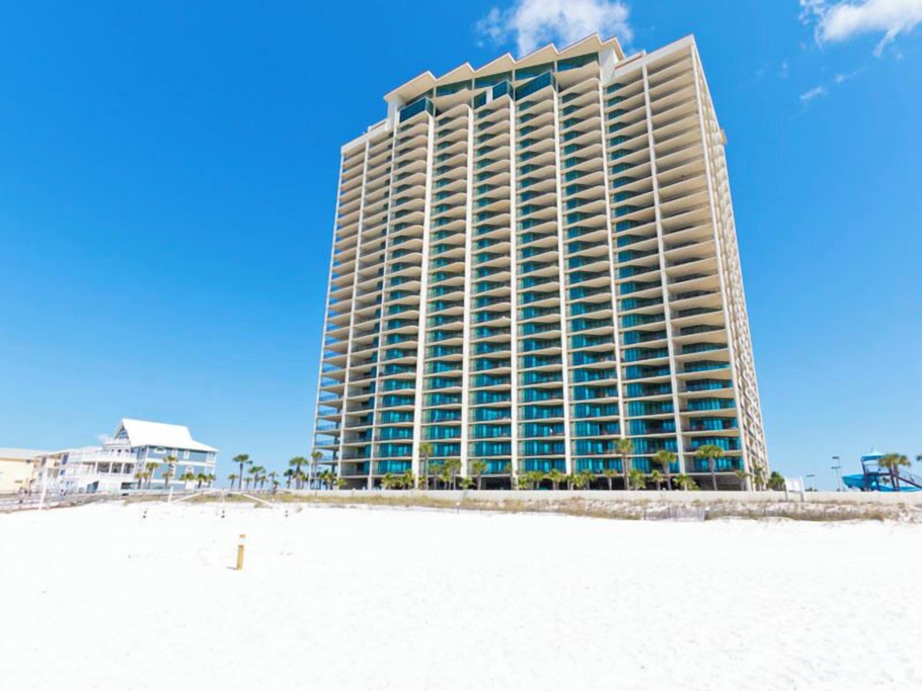 A high-rise hotel with multiple balconies, located on a beach with a clear blue sky.