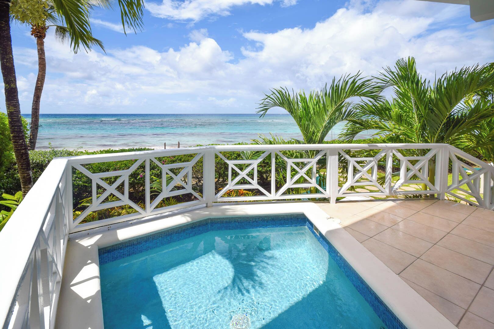 Piscina con deck blanco y barandilla, rodeada de plantas tropicales y vista a una playa.