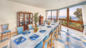 Un coin repas avec une longue table dressée pour six personnes, des chaises bleues, un buffet en bois doté de portes en verre et une vue sur la mer par de grandes fenêtres.