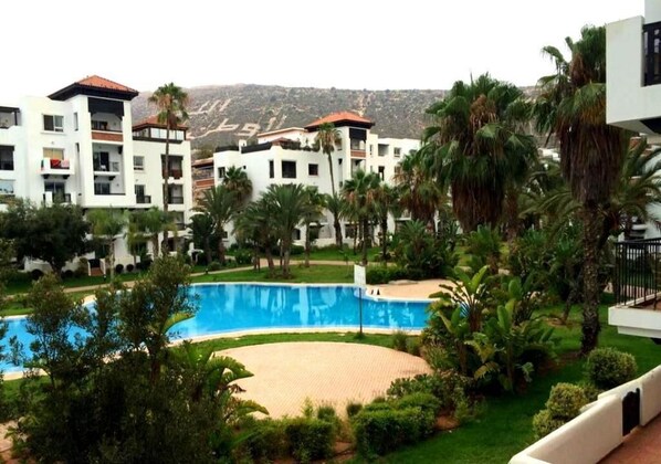 A resort with a swimming pool surrounded by palm trees and greenery, modern buildings with balconies, and a hilly backdrop.