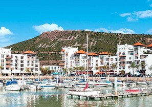 A marina with boats docked, surrounded by multi-story buildings with red roofs, and a hill in the background.