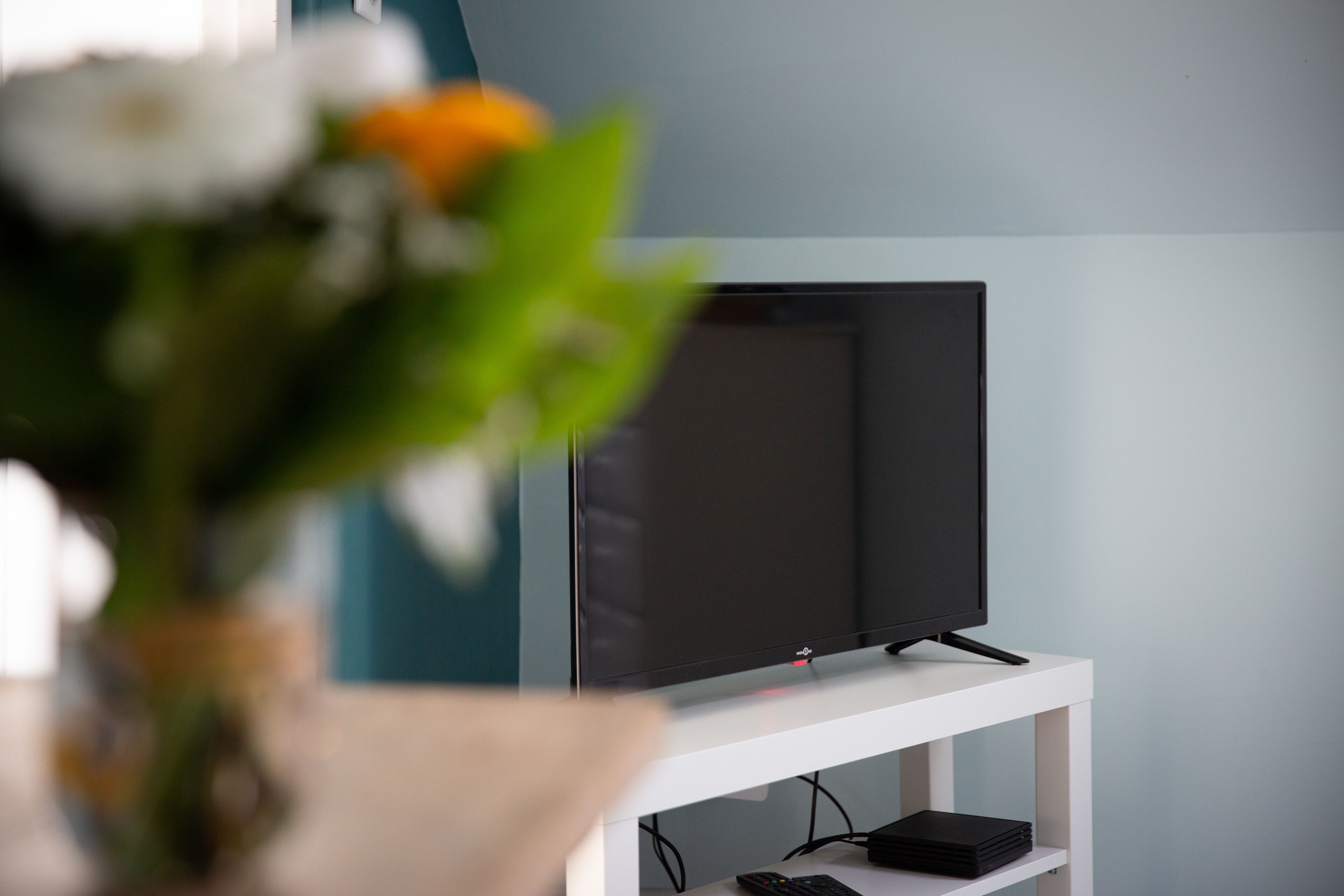 A modern living room with a flat-screen TV on a white stand, a remote control, and a black electronic device on the lower shelf.