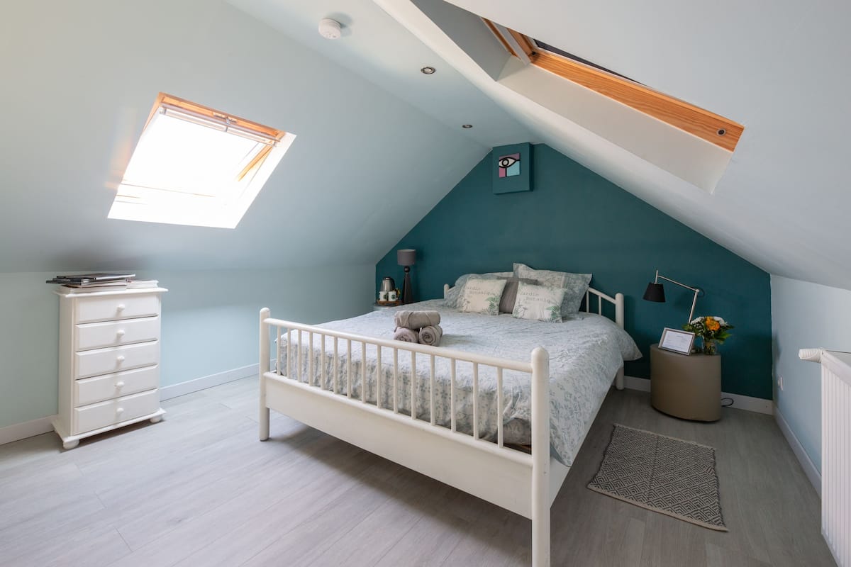 A bedroom with a sloped ceiling, a bed, a dresser, and a small table with a vase.