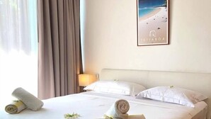 Un lit bien fait, avec du linge de lit blanc et des serviettes pliées, une table de chevet avec une lampe et, au mur, un tableau représentant une plage.