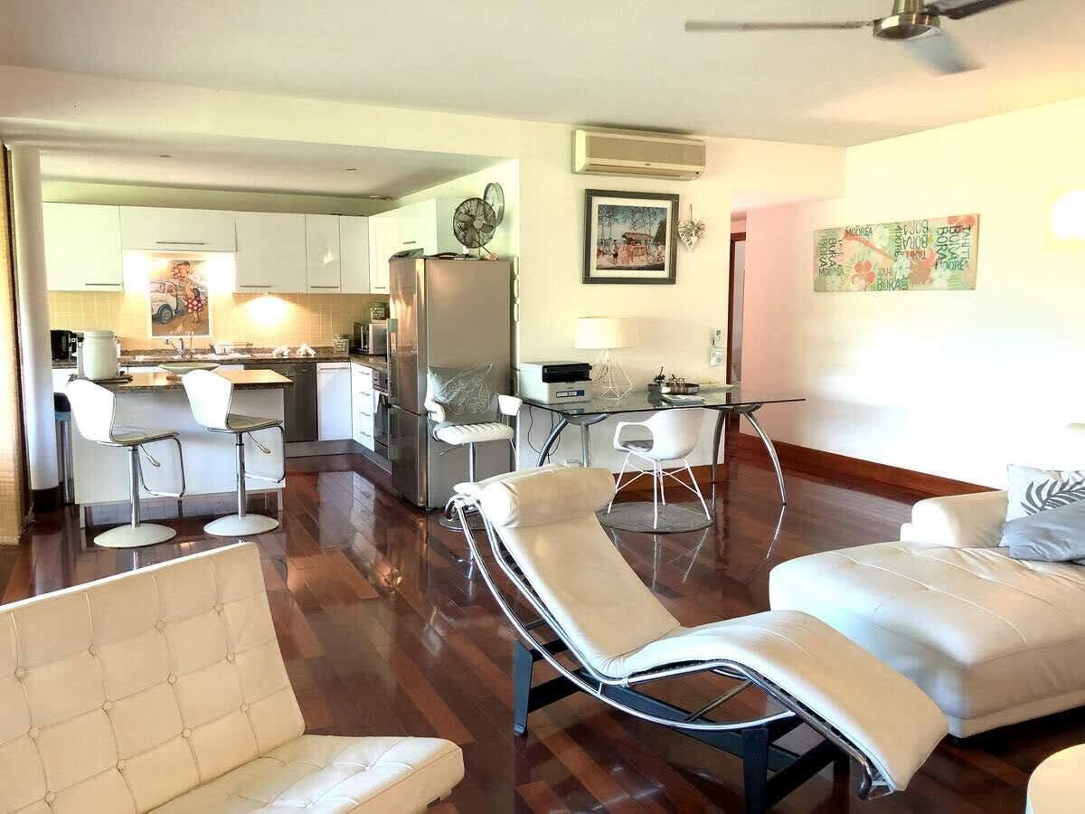 A modern living room with a sofa, a reclining chair, a dining area with a table and chairs, and a kitchen in the background.