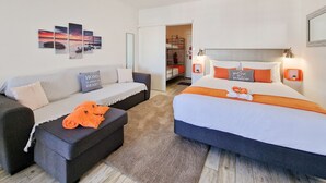 A modern hotel room with a grey sofa, a bed with orange accents, and a view of a balcony.