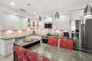 Una cocina moderna con gabinetes blancos, encimeras de granito, electrodomésticos de acero inoxidable e iluminación colgante.