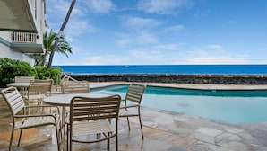 Un espace aménagé au bord de la piscine, avec des chaises et des tables, un mur en pierre et une vue sur l’océan.