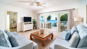 Un salon avec deux canapés gris, une table basse en bois, un téléviseur posé sur un meuble blanc et un balcon avec vue sur l’océan.