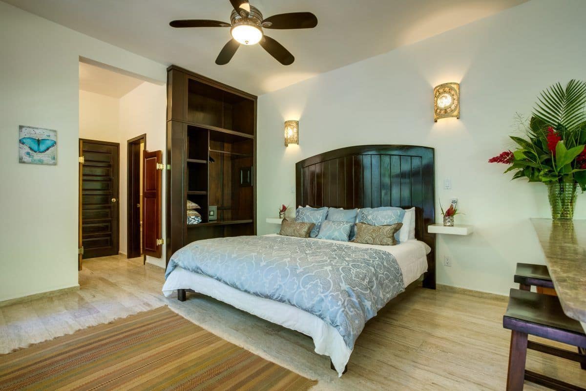 Un dormitorio con cama, ventilador de techo, mesita de noche, luz empotrada en la pared, armario de madera y puerta que da a otra habitación.
