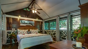 Una camera da letto con pavimento in pietra, un letto di legno, una ventola a soffitto e un'ampia finestra vista su zone verdi.