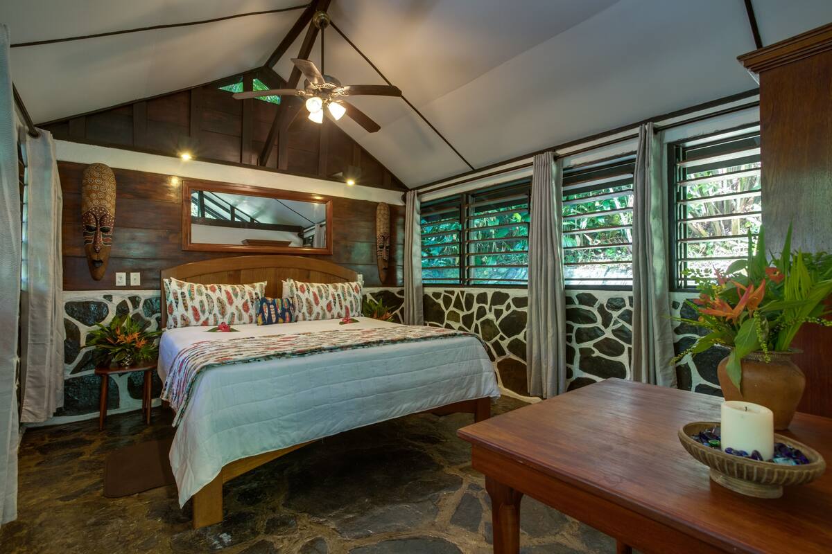 Un dormitorio con suelo de piedra, cama de madera, ventilador de techo y un amplio ventanal con vistas al verdor.