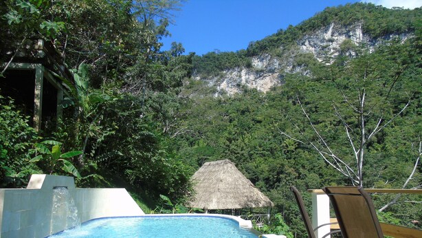 Un ponte in legno con piscina, una struttura con tetto di paglia e vista su una montagna ricoperta di bosco.