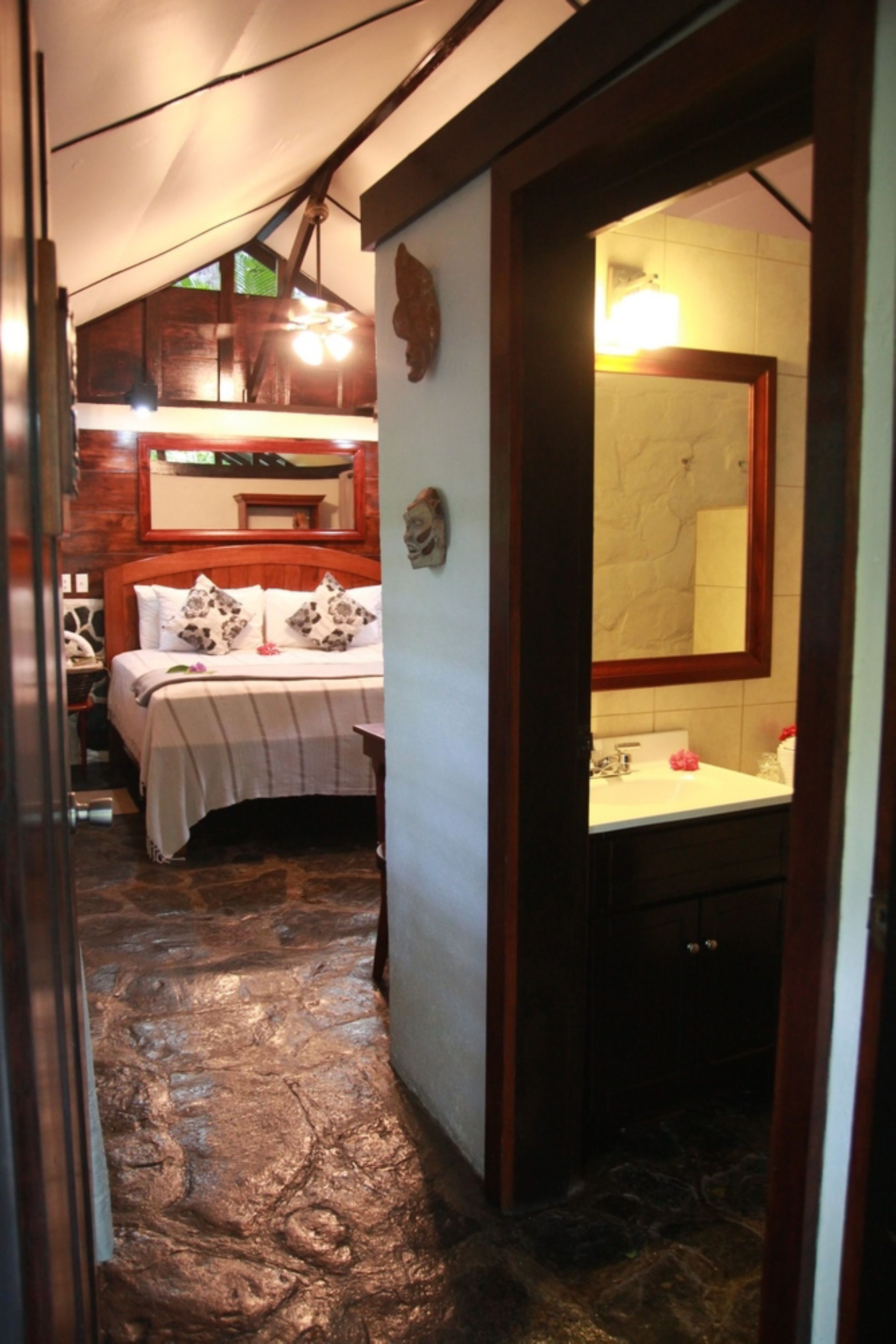 A bedroom with a bed, a wooden ceiling, a mirror, and a bathroom with a sink and toilet.