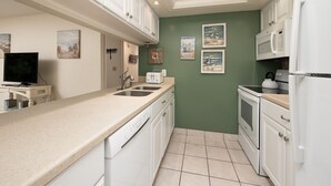 Een keuken met witte kastjes, een vaatwasser, een magnetron en een waterkoker. Er hangen prestaties aan de muur en een televisie op een standaard.