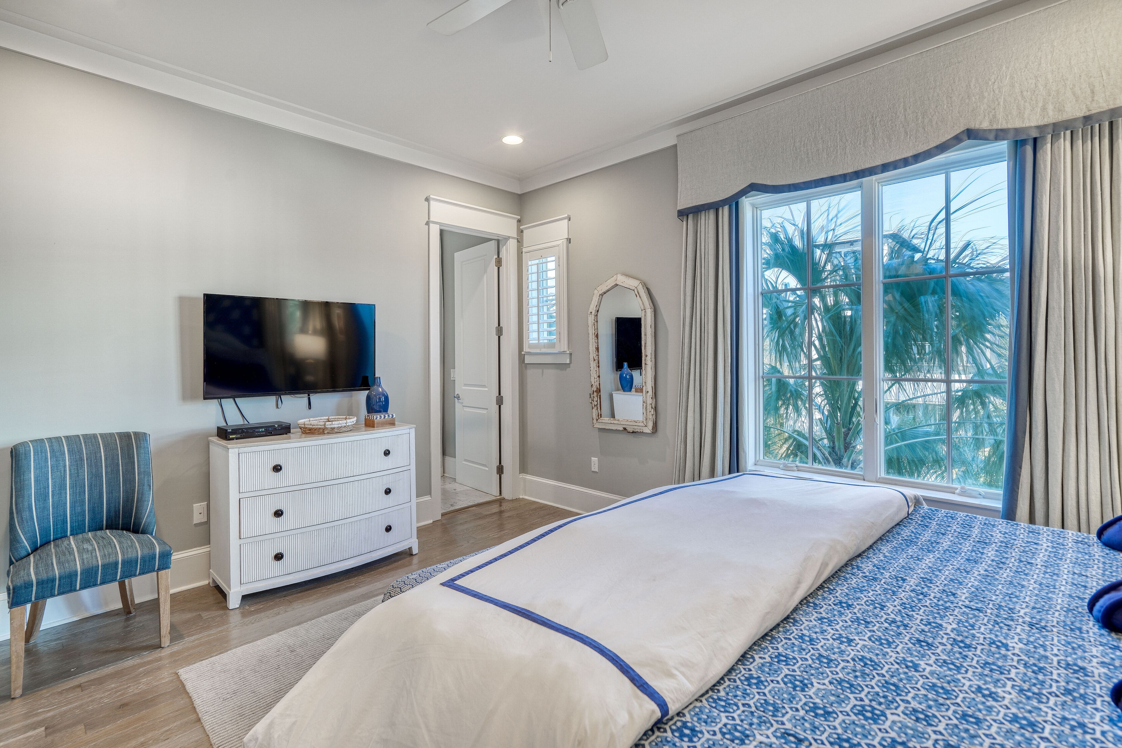 A bedroom with a large bed, a television, a dresser, and a window with a view of palm trees.