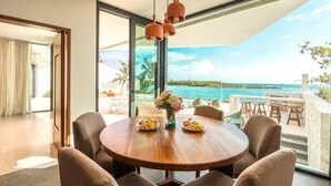A dining area with a round wooden table, beige upholstered chairs, and a view of the ocean through large windows. - Stunning Caribbean View with Entrance to Yal-ku Lagoon Akumal (Akumal)