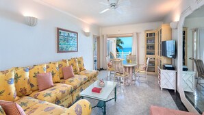 A living room with a sofa, coffee table, and a TV. There is a view of the ocean through an open door. - Private Beachfront Oasis - Reeds House 1 (3 bed) (Reeds Bay)