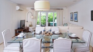 Ein Essbereich mit einem Glastisch, weißen Stühlen und einer Couch im Hintergrund.