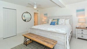 Quarto com cama, um banco, mesa de cabeceira, ventilador de teto e uma porta.